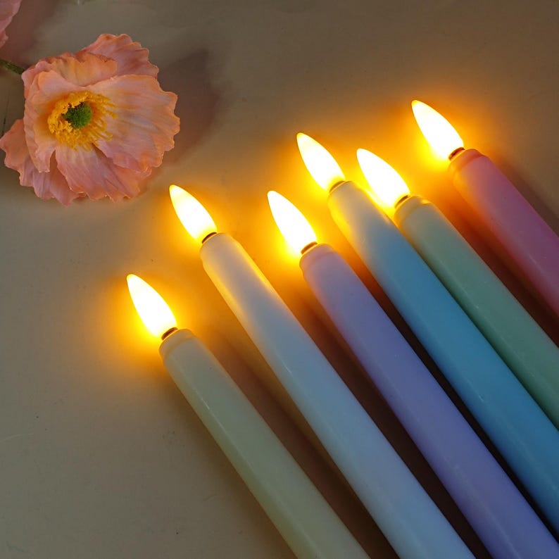 May include: A selection of pastel-coloured LED taper candles, including green, blue, purple, and pink. The candles have a realistic flickering flame effect, creating a warm ambiance. A pink flower is in the background.