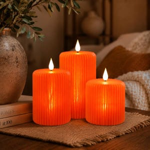 May include: Three cylindrical orange LED candles with a vertical ribbed texture. Each candle features a small, white, teardrop-shaped flame-like light at the top. The candles vary in height, with the tallest positioned in the centre.