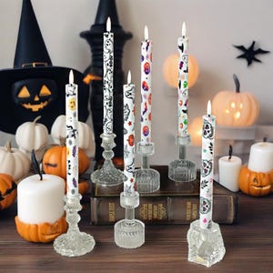 May include: A collection of Halloween-themed candles with various patterns, including ghosts, bats, and pumpkins. The candles are displayed in decorative glass holders, alongside pumpkins and a witch's hat, creating a festive atmosphere.