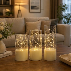 Pode incluir: Três castiçais de vidro com luzes de fadas brancas quentes. Os castiçais cilíndricos têm uma base de cor creme. As luzes são enroladas dentro do vidro, criando um brilho ambiente e acolhedor. Os castiçais estão sobre uma mesa de madeira.