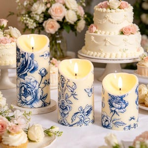 May include: Three ivory-colored pillar candles with a blue floral design and a flickering LED flame. The candles are decorated with blue roses, butterflies, and other floral elements. The candles are on a white surface, with cakes and flowers in the background.