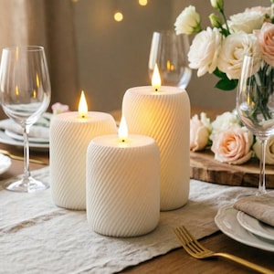 May include: Three white pillar candles with a textured surface and a warm, flickering light. The candles are on a table with wine glasses, flowers, and a gold fork. The candles have a spiral design.