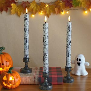 May include: Three white Halloween candles with a ghost pattern and the word "boo" are displayed on antique-style holders. A carved pumpkin and a knitted ghost are also in the image, along with autumn leaves and fairy lights.