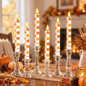 May include: Six white taper candles featuring an orange and yellow maple leaf pattern, each topped with a glowing faux flame. The candles are held in clear glass candlesticks of varying heights, arranged on a surface with pumpkins and foliage.