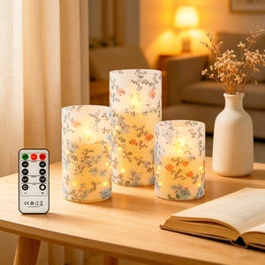 May include: Set of three floral-patterned LED candles in varying heights, with a remote control. The candles feature a white background with blue, orange, and pink floral designs. A small white vase with dried flowers and an open book are also on the table.
