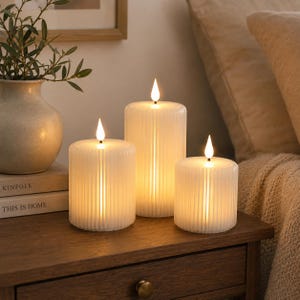 May include: Three white cylindrical LED candles with a vertical ribbed texture. Each candle features a small, glowing flame-shaped bulb at the top, emitting a warm, soft light. The candles vary in height, creating a tiered arrangement.