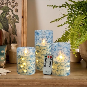 May include: Three blue and white patterned glass candle holders with flickering LED candles. The candles are on a wooden surface with a remote control for the candles.