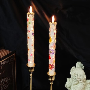 Puede incluir: Dos velas LED decorativas con diseño floral. Las velas son blancas con flores coloridas y pequeñas luces que las envuelven. Se colocan en candelabros dorados. El fondo es oscuro.