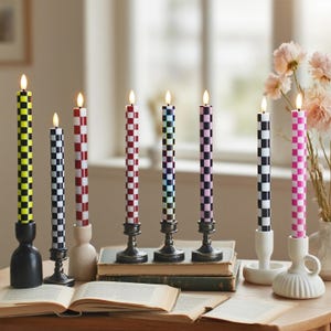 Puede incluir: Una colecci&oacute;n de velas c&oacute;nicas a cuadros en varios colores, incluyendo amarillo, negro, rojo, azul, morado y rosa. Las velas se exhiben en diferentes candelabros sobre una mesa con un libro abierto y flores.