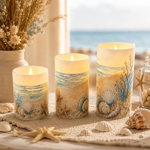 May include: Three cylindrical LED pillar candles of varying heights, each featuring a coastal scene with sandy beaches, blue ocean waves, and detailed illustrations of seashells and starfish. The candles emit a warm, soft glow from the top.