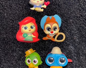 Può includere: Un set di sei piccole figurine di plastica di personaggi Disney. I personaggi includono Biancaneve, Ariel, Chip, un uccello blu, un uccello verde e un personaggio blu con un binocolo. Le figurine sono tutte colorate e hanno occhi grandi.