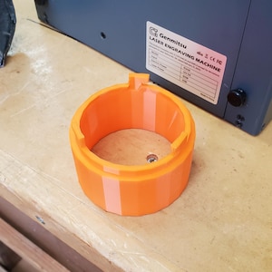 May include: Orange 3D printed ring with a smooth, rounded surface. The ring is designed to fit around a laser engraving machine.