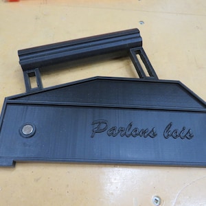 Könnte beinhalten: Schwarzes 3D-gedrucktes Holzbearbeitungswerkzeug mit Griff und dem Text "Parlons bois". Das Werkzeug hat eine rechteckige Basis mit einem kreisförmigen Magneten und einem erhöhten Rand. Der Griff ist geriffelt.