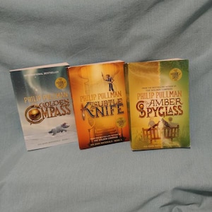 Peut inclure: Trois livres brochés d'une série intitulée "À la croisée des mondes" de Philip Pullman. Les livres sont "La Boussole d'or", "Le couteau subtil" et "Le miroir d'ambre".