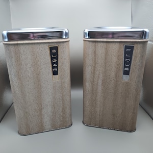 Op de afbeelding: Twee vintage keukencanners met een houtnerf afwerking. De canisters hebben een rechthoekige vorm met een zilveren metalen rand en basis. Op één canister staat "SUGAR" en op de andere "FLOUR" in een verticale letter.