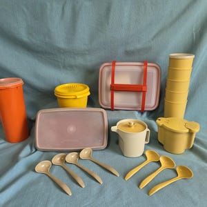 May include: A selection of vintage Tupperware containers and serving utensils in various colours. Includes an orange tumbler, yellow bowls, pink and white rectangular containers, a cream-coloured jug, and yellow and cream-coloured spoons. The items are arranged on a blue background.