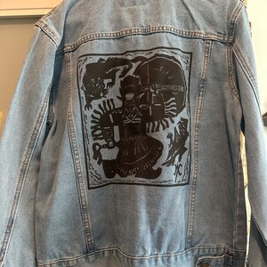May include: A blue denim jacket with a black graphic print on the back. The graphic features a stylised figure with a headdress and a sword, surrounded by other figures and symbols.