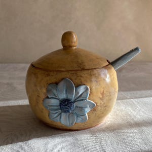 May include: A ceramic sugar bowl with a lid and spoon. The bowl is a warm yellow color with a blue flower design on the front. The spoon is light blue. The bowl is sitting on a white surface.
