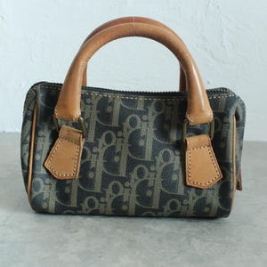 May include: A small, vintage handbag with a brown and navy blue monogram pattern. The bag features two rounded brown leather handles and matching leather accents. The bag is approximately 20 cm wide and 13 cm tall.