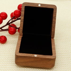 May include: Open wooden jewellery box with a black velvet interior. The box is made of light brown wood and has a square shape. A sprig of red berries is in the background.