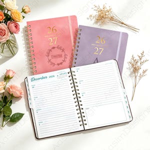 May include: Two spiral-bound planners in pink and purple with gold foil text reading 26 27, and one open planner showing a December 2026 weekly layout with lined pages, a monthly calendar, and a notes section, all featuring a white wire-o binding.