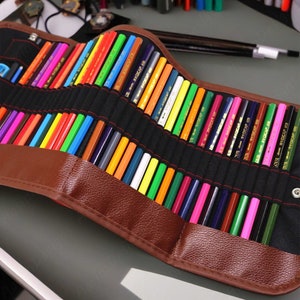 Peut inclure: Un étui à crayons en cuir marron avec une fermeture éclair. L'étui est rempli de crayons de couleur, chacun avec une étiquette dorée indiquant "Art-Scrap 48".
