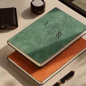 Peut inclure: Deux journaux reliés en cuir, l'un vert et l'autre orange, empilés l'un sur l'autre. Le journal vert porte les lettres "UR" et un motif floral en relief sur la couverture.