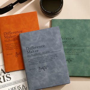 Peut inclure: Trois carnets orange, gris et vert, chacun avec le titre "Difference Maker". Les couvertures texturées incluent une définition et le nom "Beata".