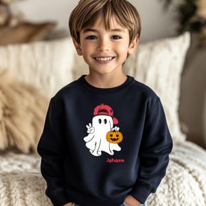 Könnte beinhalten: Marineblaues Sweatshirt mit einem Cartoon-Geist, der eine rote Baseballkappe trägt und einen Kürbis hält. Der Geist zeigt das Friedenszeichen. Der Name "Johann" ist rot darunter gedruckt. Ein Kind trägt das Sweatshirt.
