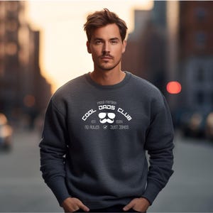 Könnte beinhalten: Ein dunkelgraues Sweatshirt mit dem weißen Schriftzug "COOL DADS CLUB", einer Sonnenbrillen- und Schnurrbart-Grafik. Der Text "EST. 2024" und "NO RULES ✓ JUST JOKES" sind ebenfalls zu sehen. Das Sweatshirt hat einen Rundhalsausschnitt.