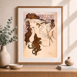 May include: A framed art print featuring an Alphonse Mucha illustration of a woman with long, flowing brown hair, wearing a sheer veil and holding a sprig of white flowers. The text SALON DES CENT is visible at the top in a stylised font.