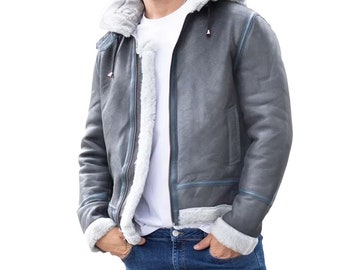 Può includere: Una giacca di pelle di pecora grigia con cappuccio e chiusura a zip. La giacca ha una fodera in pelle di pecora bianca e un bordo blu.