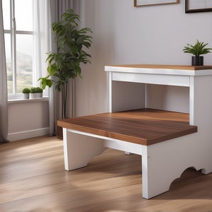 Puede incluir: Taburete de madera blanco con dos peldaños y un estante. El peldaño superior es de madera y el inferior es blanco. El estante está abierto y tiene un marco blanco.