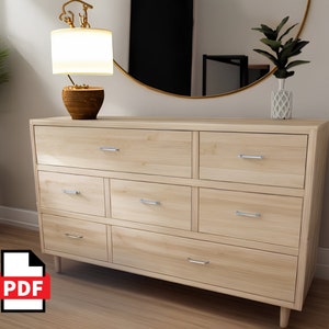 Könnte beinhalten: Eine Kommode aus hellem Holz mit sechs Schubladen und silbernen Schubladengriffen. Die Kommode hat ein modernes Design mit leicht angeschrägten Beinen. Die Kommode steht in einem Raum mit einem Spiegel und einer Lampe auf einem Beistelltisch.