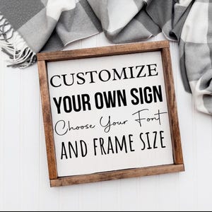 以下が含まれることがあります： 「CUSTOMIZE YOUR OWN SIGN Choose Your Font AND FRAME SIZE」の文字が黒で書かれた、額縁に入った木製の看板。看板は白い背景と濃い茶色の木製フレームです。グレーと白のチェック柄の毛布が背景にあります。