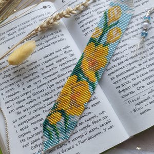Könnte beinhalten: Ein langes, flaches, gewebtes Perlen-Lesezeichen mit einem Muster aus gelben Tulpen mit grünen Stielen und Blättern auf hellblauem und weißem Grund. Die Enden sind mit hellem Garn, kleinen Metallperlen und blauen Glasperlen verziert.