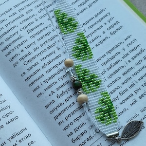 Puede incluir: Un marcapáginas tejido con fondo blanco y un patrón repetitivo de tréboles verdes. La parte superior está decorada con una cuenta verde oliva, mientras que la inferior incluye un dije de hoja de metal plateado y varias cuentas de madera y oscuras.