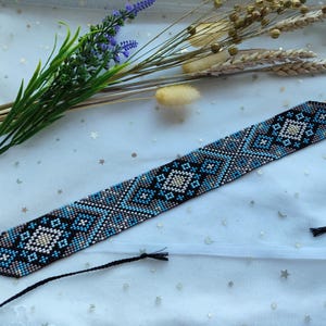 May include: A wide woven beadwork bracelet featuring a geometric diamond pattern in shades of blue, black, and cream. The band is constructed from small, uniform beads and includes thin black cord ties at both ends for adjustable fastening.