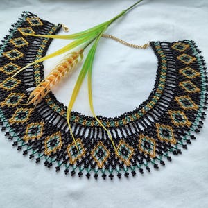 May include: A black, gold, and green beaded necklace with a geometric pattern. The necklace is in the shape of a collar and has a fringe.