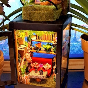 Könnte beinhalten: Eine Miniatur-Hausszene in einer schwarzen Laterne mit einer roten Couch, einem kleinen Tisch und einem Bücherregal. Das Haus ist mit Pflanzen und Büchern dekoriert. Die Laterne hat eine Glasfront und einen Metallrahmen.