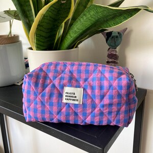 Peut inclure: Une trousse rectangulaire matelassée avec un motif vichy rose et bleu. Une étiquette en tissu blanc est cousue sur le devant, affichant le texte FELICITÀ BONHEUR HAPPINESS en lettres capitales noires sans empattement.