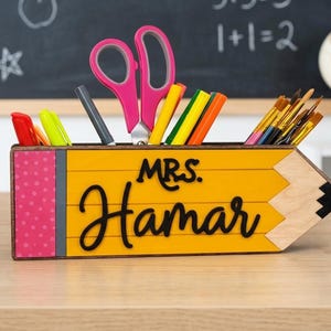 May include: A yellow pencil-shaped desk organizer with the name "Mrs. Hamar" in black script. The organizer holds pink scissors, pens, markers, and paintbrushes. The left side has a pink polka dot design.
