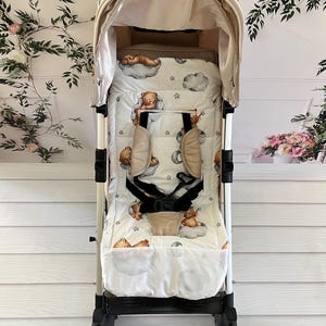 May include: A beige stroller with a white and brown patterned liner featuring teddy bears and clouds. The stroller has a black handle and black wheels.
