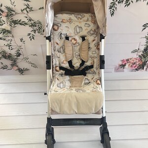 May include: Beige stroller with a cream and brown patterned liner featuring clouds, stars, and animal illustrations. The stroller has a black handle and black wheels.