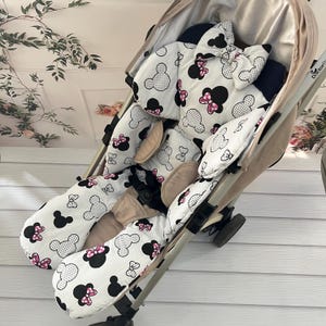 May include: A beige stroller with a white and black patterned seat cover featuring Mickey Mouse silhouettes and pink bows. The stroller has a black safety harness and is designed for infant use.