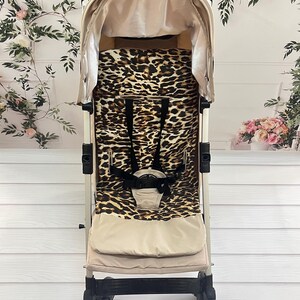 以下が含まれることがあります： ベージュとヒョウ柄のベビーカー、キャノピー付き。ベビーカーはベージュのフレーム、黒いハンドル、ヒョウ柄のシートを備えています。ベビーカーは乳幼児向けに設計されています。