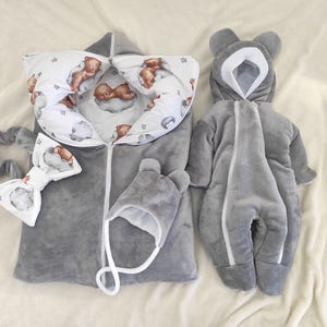 Puede incluir: Un saco de dormir para bebé gris con estampado de osos blancos y marrones, un mono con capucha gris a juego, un gorro gris con orejas de oso y un lazo blanco y gris.