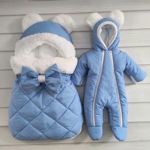 Puede incluir: Un saco para bebé y un mono de nieve a juego en azul claro. El saco tiene un diseño acolchado, un lazo grande y un interior y ribete blancos y esponjosos. El mono tiene una capucha con orejas de oso.