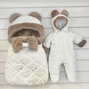 May include: A white baby bunting bag and snowsuit set. The bunting bag features a quilted pattern with gold stars, faux fur trim, and bear ears. The snowsuit is white with gold stars, a hood, and faux fur trim on the cuffs.