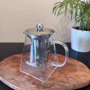 May include: A clear glass teapot with a stainless steel lid and infuser. The teapot has a handle and is marked with measurements in millilitres. The brand name "AesaCi" is printed on the front. The teapot sits on a wooden serving tray.